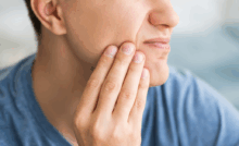 A person in a blue shirt is touching their cheek with a pained expression, indicating discomfort, possibly from jaw pain.