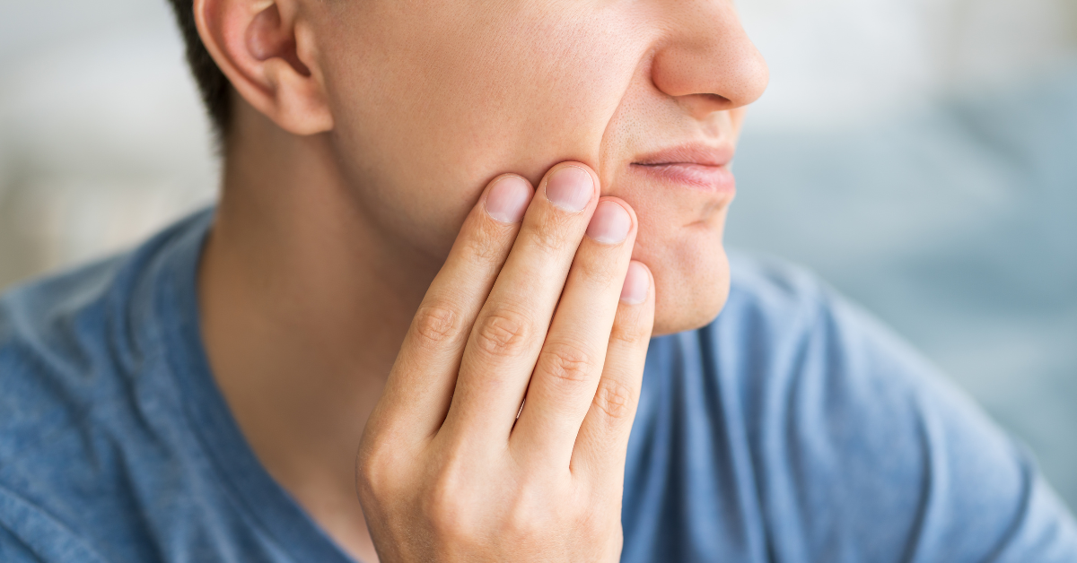 A person in a blue shirt is touching their cheek with a pained expression, indicating discomfort, possibly from jaw pain.