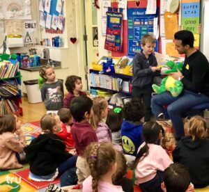A classroom of young children attentively watch a teacher holding a green dinosaur puppet. A boy interacts with the puppet, creating a lively, engaging atmosphere.