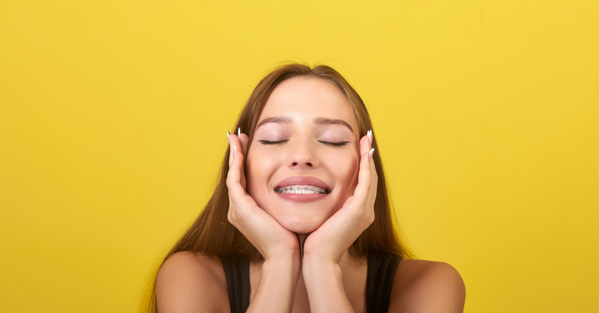 A smiling person with braces gently holds their cheeks, eyes closed, in front of a bright yellow background, conveying joy and contentment.