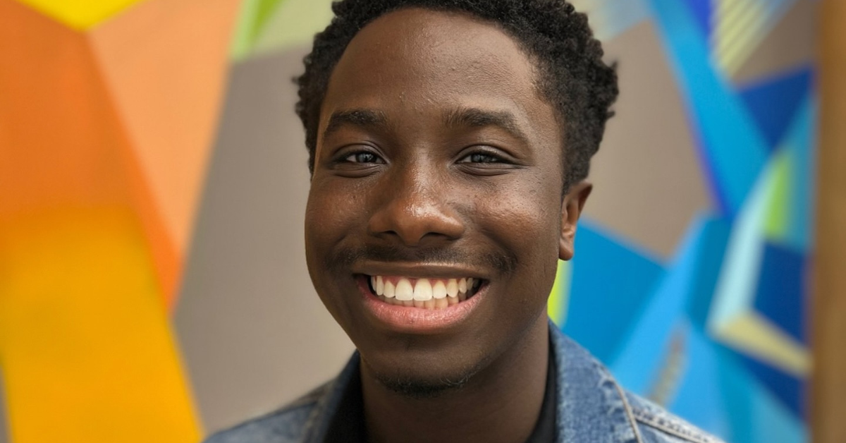 Smiling person in front of a vibrant, geometric mural with bold orange, blue, and green shapes. The joyful expression conveys a cheerful tone.
