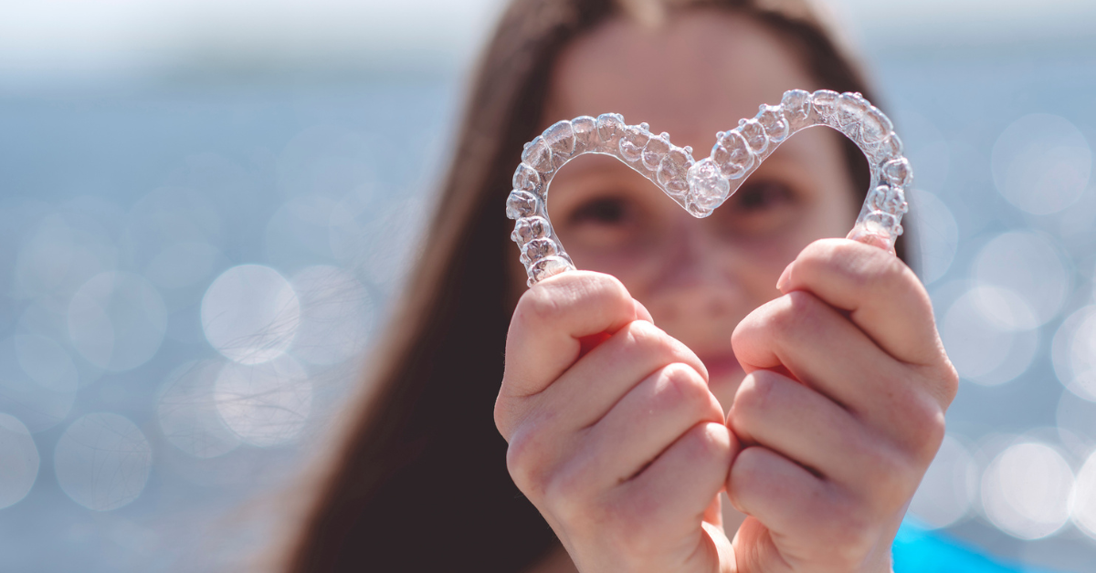 A person holds a clear, heart-shaped object made of beads against a blurred ocean background. The sun creates a shimmering, serene effect.